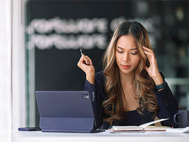 A business woman sits at her desk with a tablet while taking notes with her pen on a notepad.